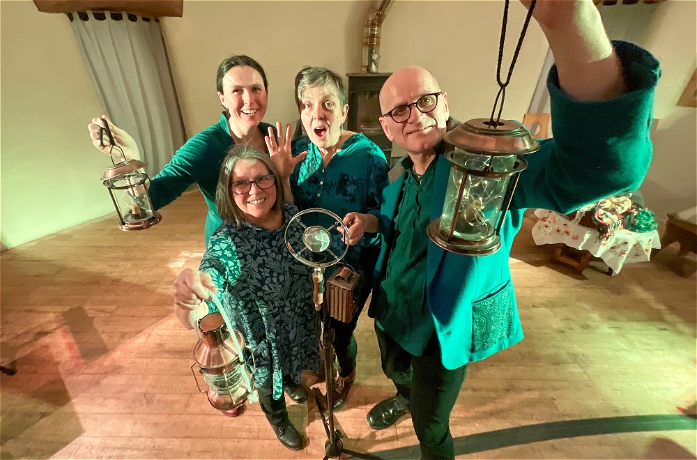 Four smiling white adults stand indoors on a wooden floor, holding lanterns towards the camera. They are gathered closely together, looking up, wearing blue-green patterned clothing, and appear cheerful and engaged.