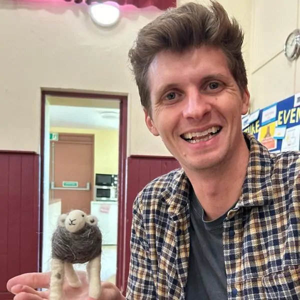 A white man in his thirties is smiling to camera. he has a big quiff and blonde hair. He is wearing a brown check shirrt and is holding a needle felted herdwick sheep.