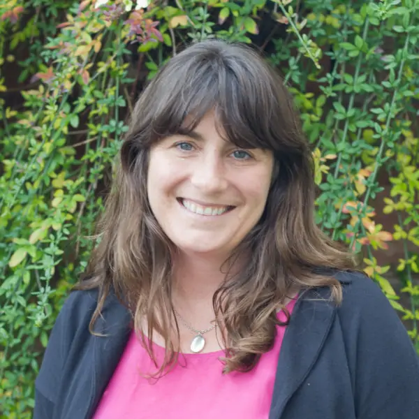 A white woman in her forties is smiling towards the camera. She has long brown hair which falls on her shoulders. She is wearing a navy jacket with. abright pink top.
