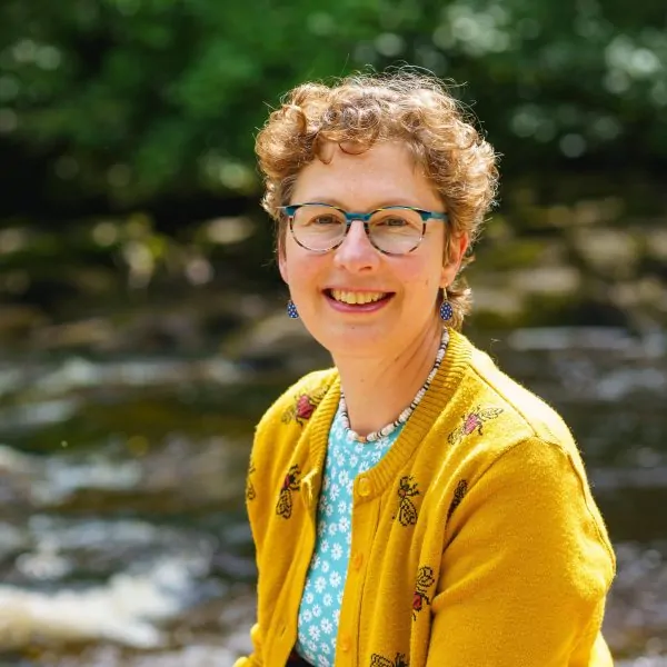 A white woman in her forties is smiling towards the camera. She has short, curly, brown hair. She is wearing glasses, a blue polkadot top and a knitted yellow cardigan.