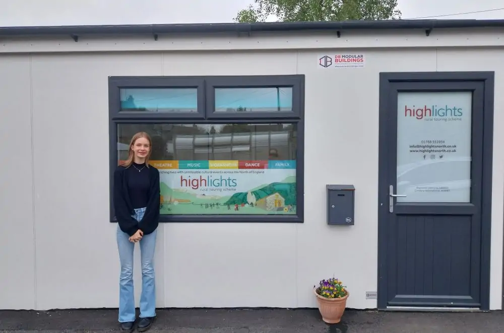 A young woman stands in front of the Highlights office. She is wearing blue jeans and a black long sleeve top. She has blonde hair and is smiling. The building is a single story portacabin. There is a decal in the window with an illustration and Highlights branding.