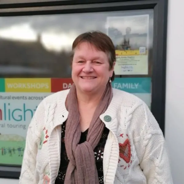 A white woman in her fifties is smiling towards the camera. She has short brown hair, and is wearing a knitted cardigan and brown scarf.