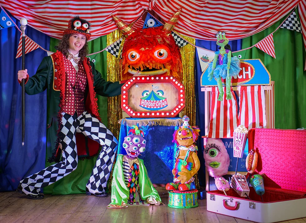 A young woman stands on a brightly decorated stage. She is wearing checkerboard trousers and a navy blue tail coat, with a polka dot waistcoat and top hat. The top hat has a googly eyes and monster teeth. On the stage with her are an array of monster puppets. They have googly eys, big grins and monster teeth.