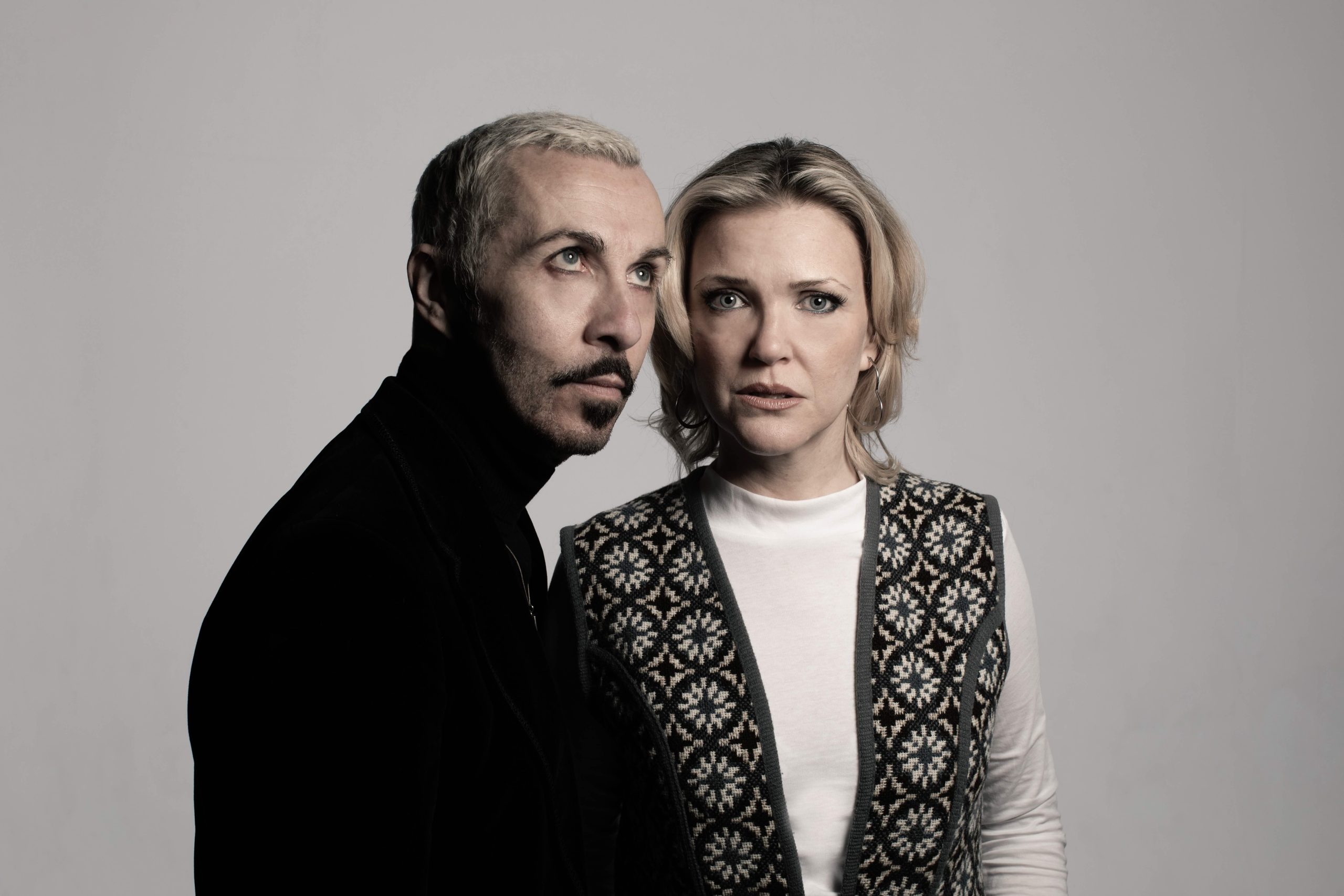 AE19A2205 (c) Andy Hollingworth Archive copy 2 A man and a woman stand side by side. He has short blonde hair, a black moustache and soul patch, and he is wearing a black roll neck. The woman has medium length blonde hair with a center parting. She wears a white top with a fairisle patterned sleeveless cardigan on top. THe man looks off to the top right of the picture, while the woman looks out towards the camera with a confused look on her face.