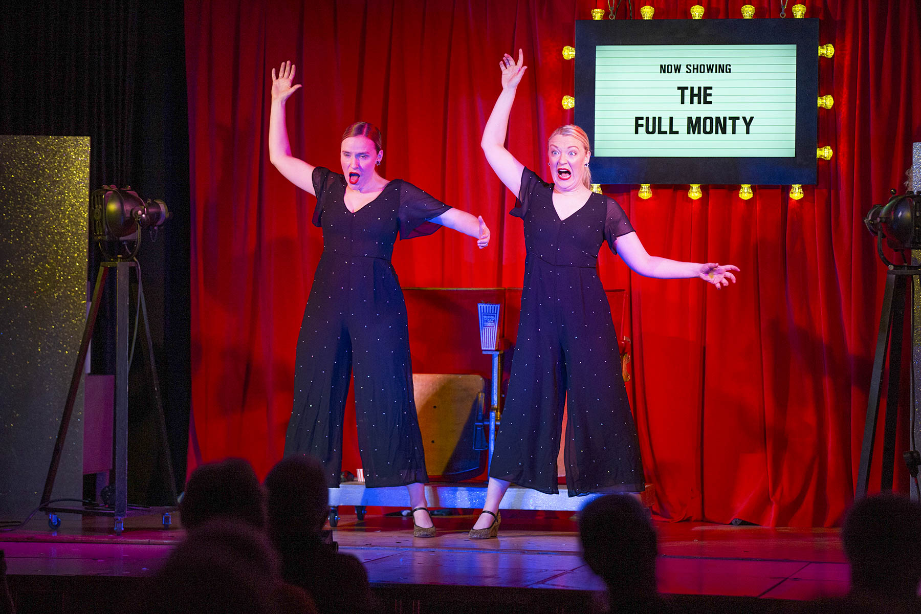 Two female performers stand on stage in front of a red curtain. There is a sign, surrounded by illuminated lightbulbs which reads 'Now Showing: The Full Monty'. The two performers have blonde hair and wear glittering jump suits. They have their right hands in the air above their heads and their left hands held out to their sides. They are pulling dramatic faces.