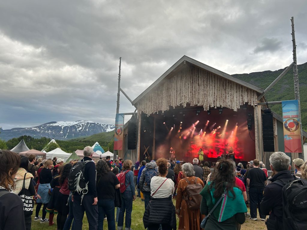 Outdoor music festival with a wooden stage, colourful lights, and performers under a cloudy sky. The audience is surrounded by tents and a scenic mountain backdrop.
