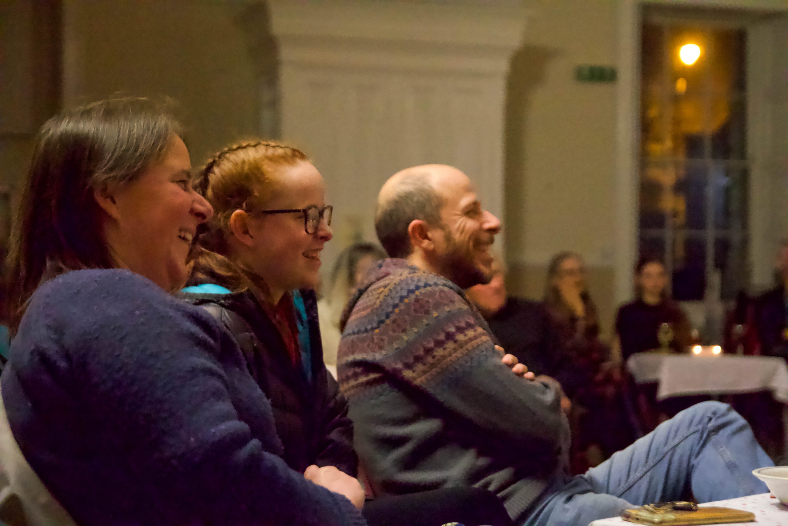 In a brightly lit hall, three audience members are seen in profile. They are all smiling and laughing.