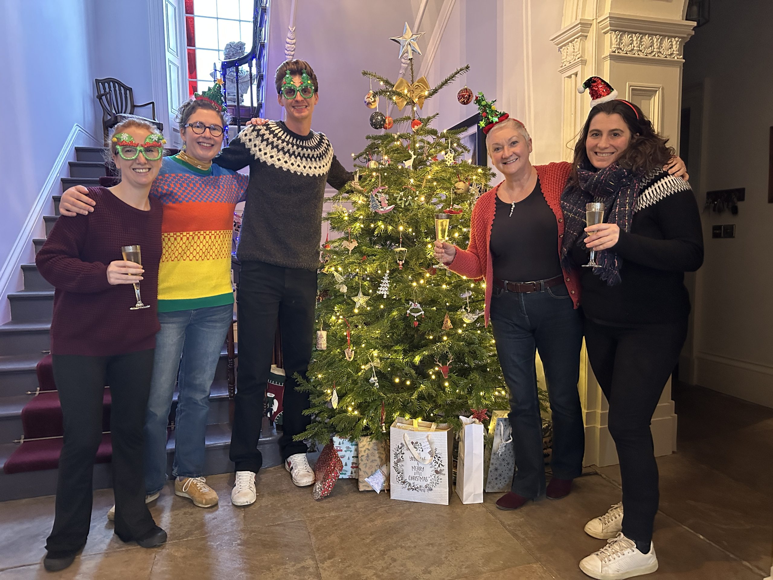 The Highlights team stand in front of a christmas tree in the hallway of a country house. There are four women of medium height, and one tall man. They are wearing christmas hats and masks.
