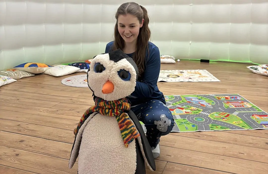 A young white woman is sat inside a giant inflatable igloo. She is crouched down behind a stuffed toy in the shape of a penguin. The woman is wearing leggings printed with a koala bear pattern, a navy top. She has dark hair tied back in two pigtails.