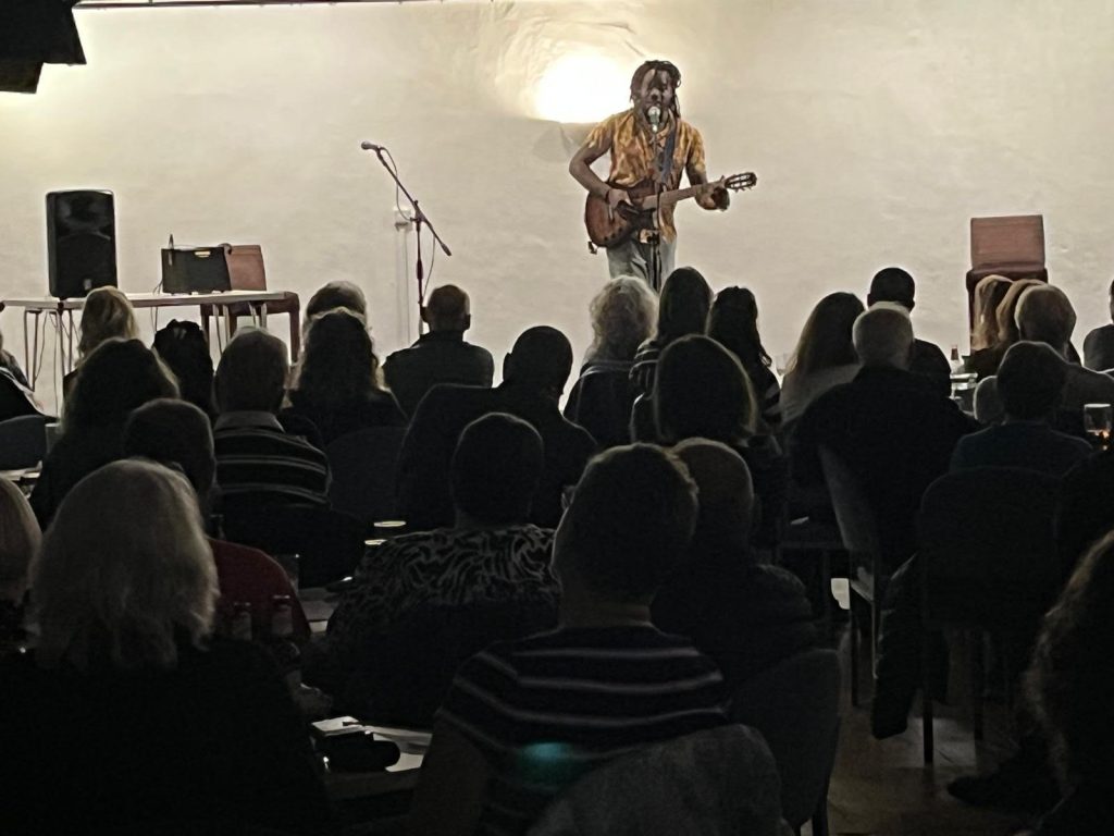 A black musician with dreadlocks plays guitar on stage in a small venue, facing a seated audience. The atmosphere is intimate and focused, with people engaged in the performance.