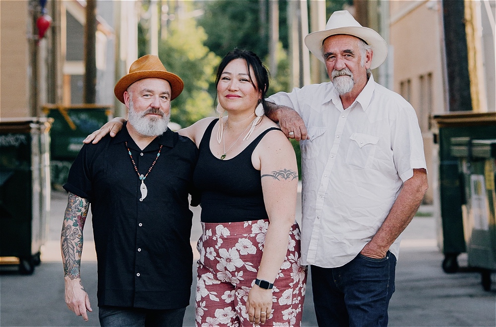 Three people stand in a street with arms over each others shoulders. The man on the left wears a black shirt, a beaded necklace, and a tan hat. The woman in the middle wears a black tank top and floral trousers. The man on the right wears a white shirt and a white hat.
