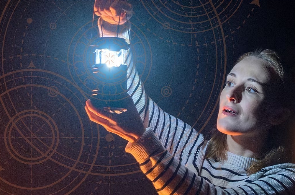 A woman holds up a lit lantern with a bright light shining from it, against a dark background with glowing circular celestial charts.