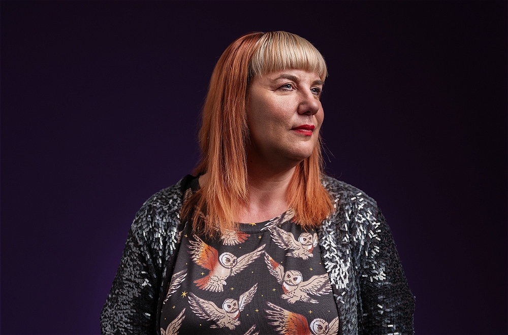A woman with red hair and a blonde fringe, wearing a sequined jacket over a dark shirt with an owl print, looks to the right against a purple background.